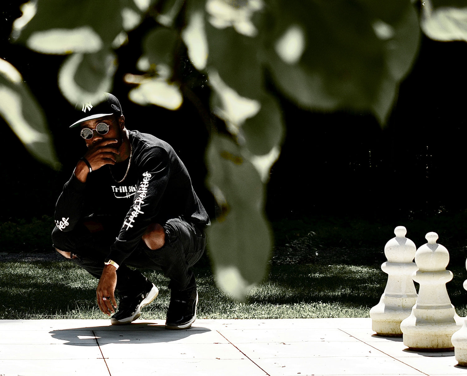 Person in black outfit with white text, crouching outdoors near large chess pieces.shop_this_look_AVdw3f